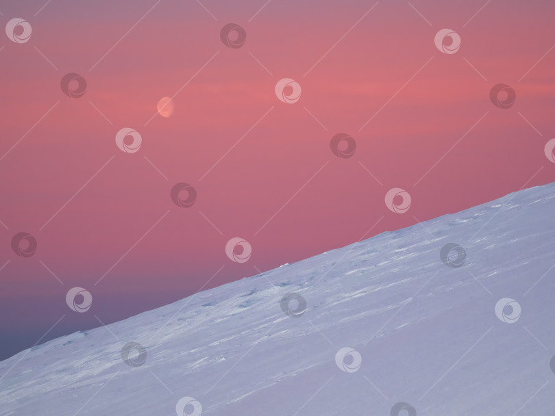 Скачать Атмосферный минималистский пейзаж с луной над высоким заснеженным горным склоном на рассвете. Розовое небо полярной зари с луной. фотосток Ozero