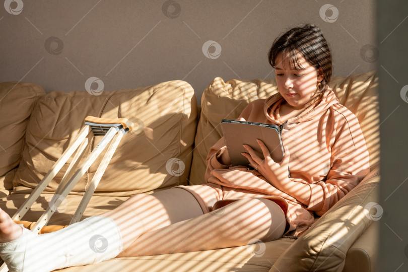 Скачать Девушка с гипсом на ноге, сидит на диване и рисует на графическом планшете. Забинтованный гипс на ноге и пальцах ног после несчастного случая с травмой при беге.Девочка-подросток в гипсе. фотосток Ozero