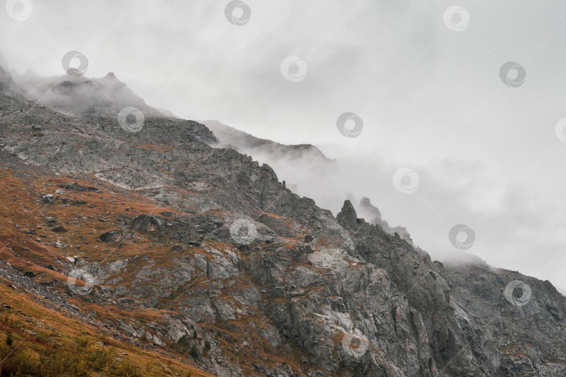 Скачать Атмосферный туманный пейзаж с нечеткими силуэтами острых скал в низких облаках во время дождя. Впечатляющий вид на большие горы, размытые дождевой дымкой в серых низких облаках. фотосток Ozero