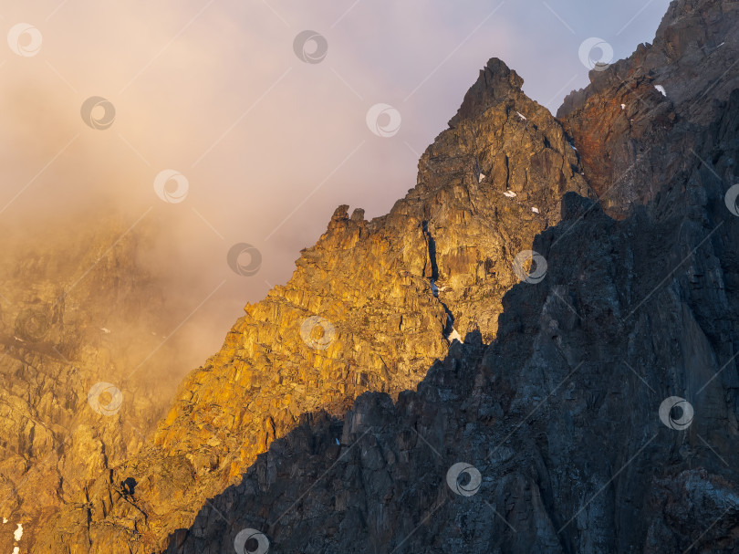 Скачать Живописный пейзаж с золотистым светом на вершине горы в низких облаках в желтых тонах восхода солнца. Красочный вид на горы и скалы в лучах утреннего солнца сквозь низкие облака. Яркий пейзаж с золотистыми скалами. фотосток Ozero
