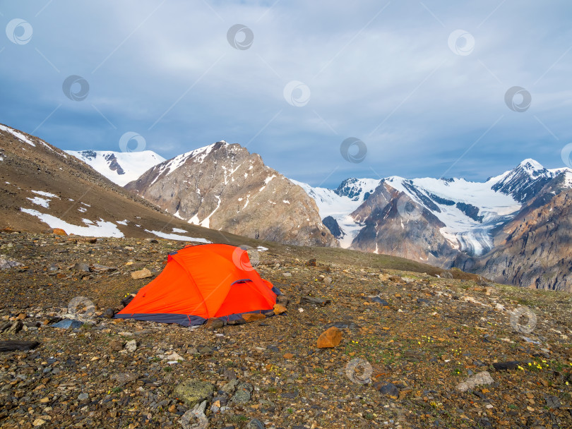 Скачать Летний кемпинг в горах. Яркий альпийский пейзаж с ярко-оранжевым шатром на очень большой высоте с видом на высокую гору и большой ледник в темных облаках. Потрясающий горный пейзаж с палаткой. фотосток Ozero