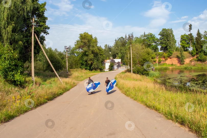 Скачать Две молодые женщины несут SUP-доски к воде. Деревенский летний пейзаж, активный отдых. фотосток Ozero