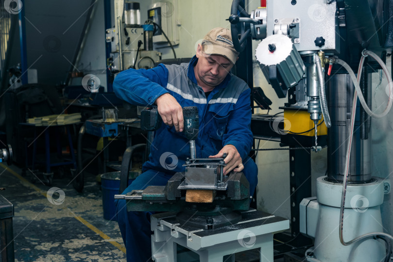 Скачать рабочий в полутемной мастерской закрепляет несколько заготовок в стопке с помощью электрической отвертки фотосток Ozero