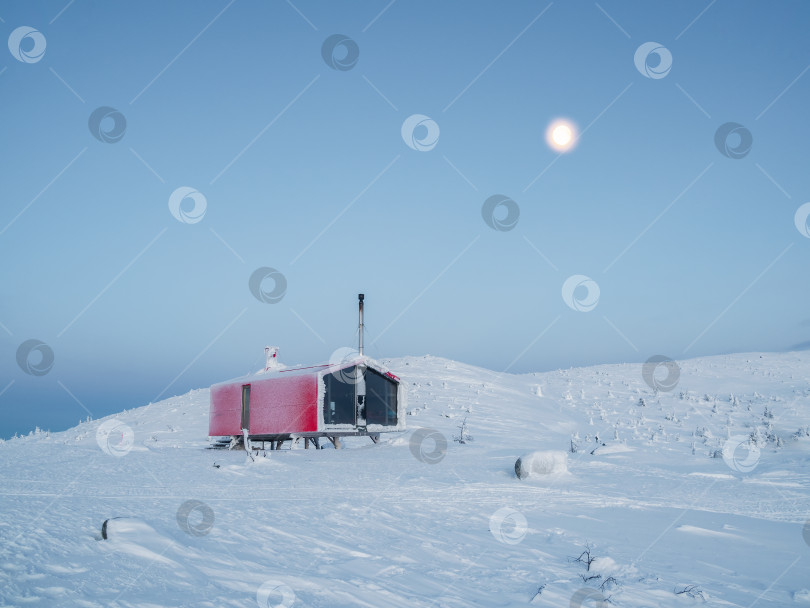 Скачать Одинокое красное бунгало на заснеженном морозном холме под темным вечерним небом с полной луной. Минималистичный фон с изображением одинокой красной хижины зимой. фотосток Ozero