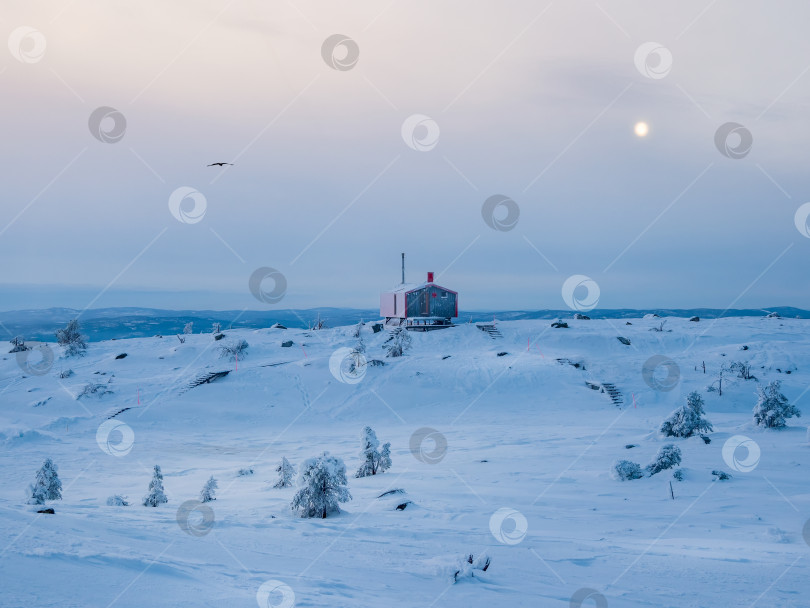 Скачать Одинокое красное бунгало на заснеженном морозном холме под темным вечерним небом с полной луной. Минималистичный фон с изображением одинокой красной хижины зимой. фотосток Ozero