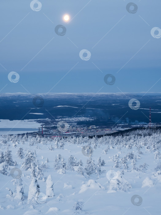 Скачать Полная луна над заснеженным склоном полярной сопки зимой с отдаленным видом на Кандалакшу, портовый город на севере России. Зимний полярный пейзаж. Холодная зимняя погода. Суровый северный климат фотосток Ozero