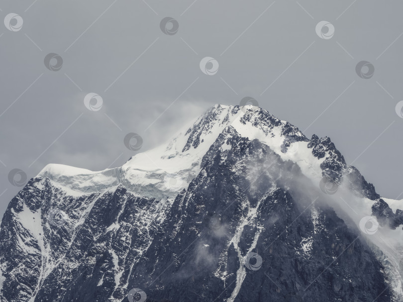Скачать Атмосферный альпийский пейзаж с заснеженной горной вершиной под снежно-серым небом. Потрясающий пейзаж с красивым остроконечным пиком, покрытым снегом, и высокой снежной горной стеной с низкими облаками. фотосток Ozero
