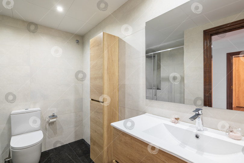 Скачать Современная ванная комната со стенами, выложенными белой плиткой. Небольшой деревянный шкафчик для туалетных принадлежностей и полотенец расположен у стены и гармонирует по цвету со шкафчиком для раковины. Зеркало отражает дверной проем в комнату. фотосток Ozero