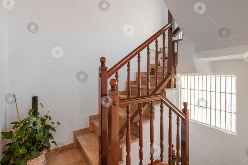 Скачать Просторная лестница с мраморными ступенями и деревянными перилами, ведущая на верхний этаж на фоне белых стен. Яркий дневной свет из окна хорошо освещает лестницу и растение в горшке на полу. фотосток Ozero
