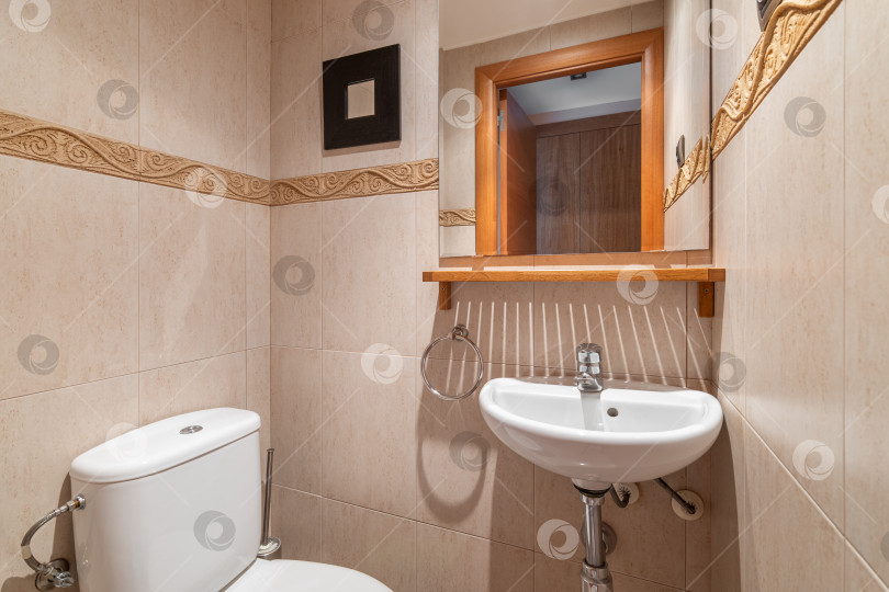 Скачать Небольшая ванная комната с туалетом и умывальником, зеркалом на стене, в котором видна открытая дверь. Стены комнаты отделаны керамической плиткой красивого нежного цвета с яркой полосой орнамента. фотосток Ozero
