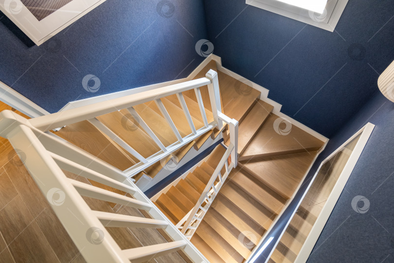 Скачать Вид сверху на светлую винтовую лестницу с полированными деревянными паркетными ступенями и матово-белыми перилами на фоне голубых стен. Дневной свет, льющийся из окна, освещает винтовую деревянную лестницу. фотосток Ozero