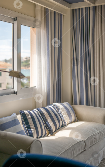 Скачать Крупным планом уютный мягкий диван бежевого цвета с декоративными подушками в вертикальную сине-бежевую полоску и занавесками в том же стиле. Уютная гостиная, залитая ярким солнечным светом из окна. фотосток Ozero