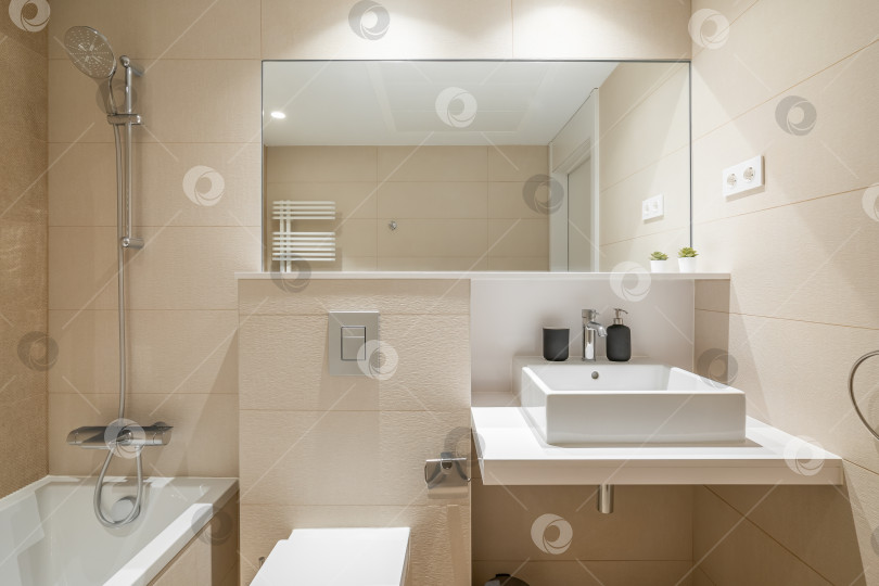 Скачать Уютная ванная комната в современном дизайне с огромным зеркалом на стене и ярким освещением. Стены отделаны бежевой плиткой и белой сантехникой. Комната для принятия утреннего душа и расслабляющих вечерних ванн. фотосток Ozero