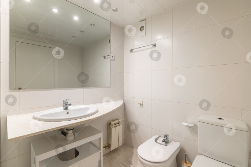 Скачать Простая белая ванная комната с большим зеркалом, классической раковиной, биде и туалетом. фотосток Ozero