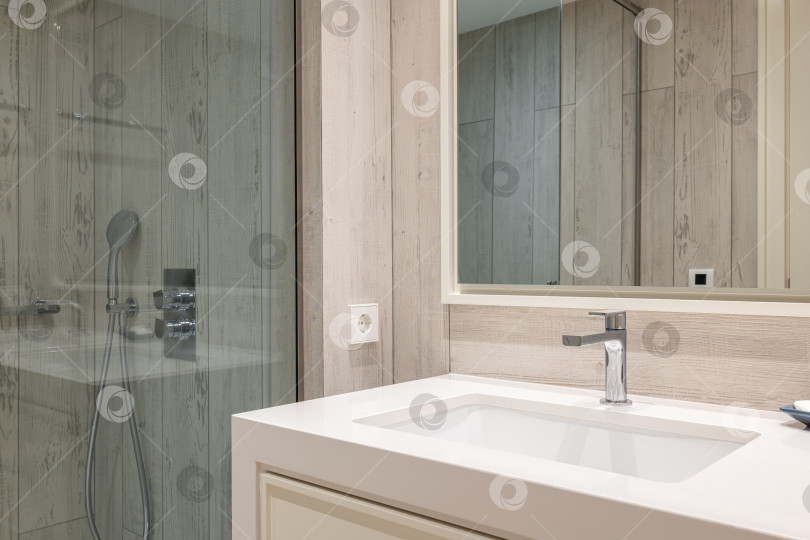 Скачать Светлая ванная комната крупным планом с панельными стенами с эффектом дерева, квадратной керамической раковиной с краном на мраморной столешнице и зеркалом на стене. Душевая зона огорожена стеклянной стеной голубоватого оттенка. фотосток Ozero