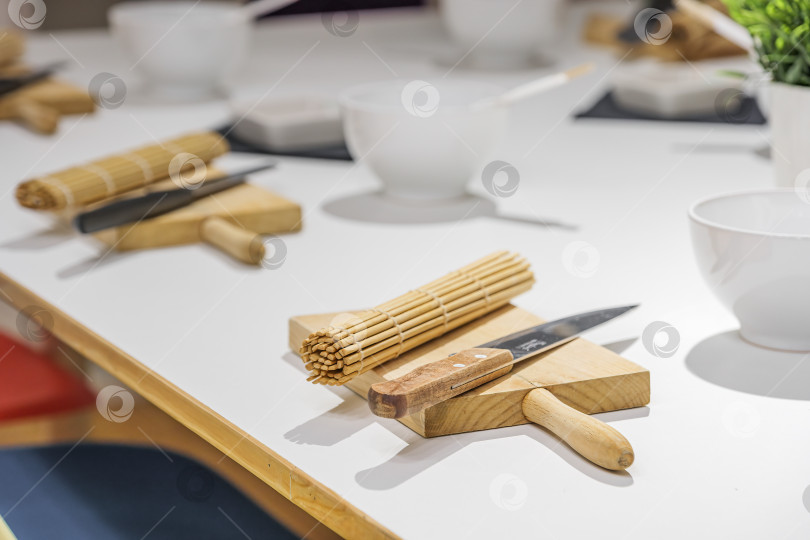 Скачать Наборы ножей, доски и бамбукового коврика для суши на белом столе перед мастер-классом по приготовлению суши. Концепция образования, кулинарии и развлечений. фотосток Ozero
