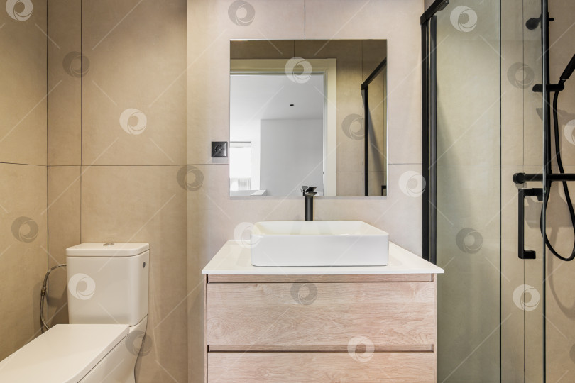 Скачать Современная ванная комната выложена бежевой плиткой. Квадратное зеркало, унитаз, белая раковина и душевая зона со стеклянной перегородкой. фотосток Ozero