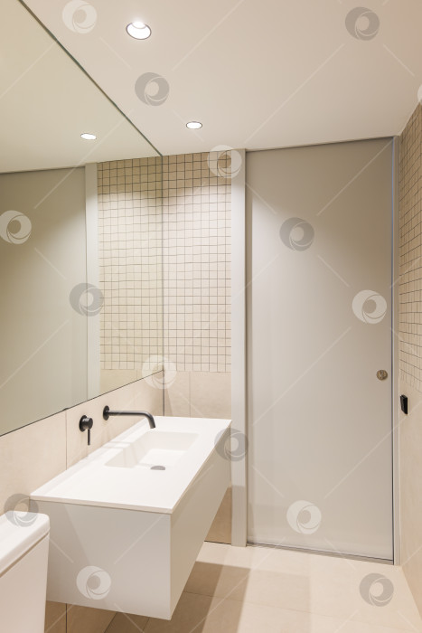 Скачать Вертикальный вид узкой ванной комнаты в стиле минимализма. Большое зеркало и белая раковина с черным краном. фотосток Ozero