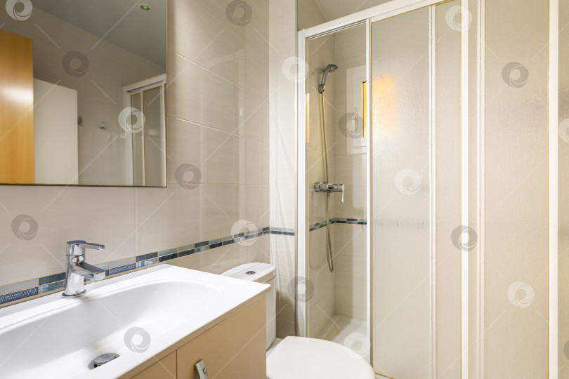 Скачать Интерьер современной ванной комнаты, выложенной плиткой, с белой раковиной, деревянной мебелью, зеркалом и душевой кабиной. фотосток Ozero