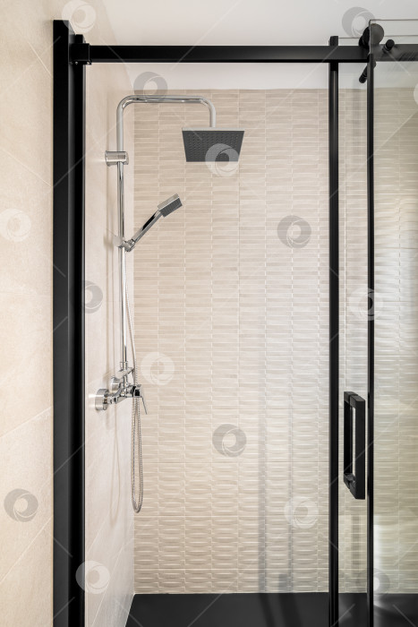 Скачать Современная ванная комната, отделанная кафелем, с дождевой насадкой, ручным душем и стеклянной дверью с черным обрамлением. фотосток Ozero