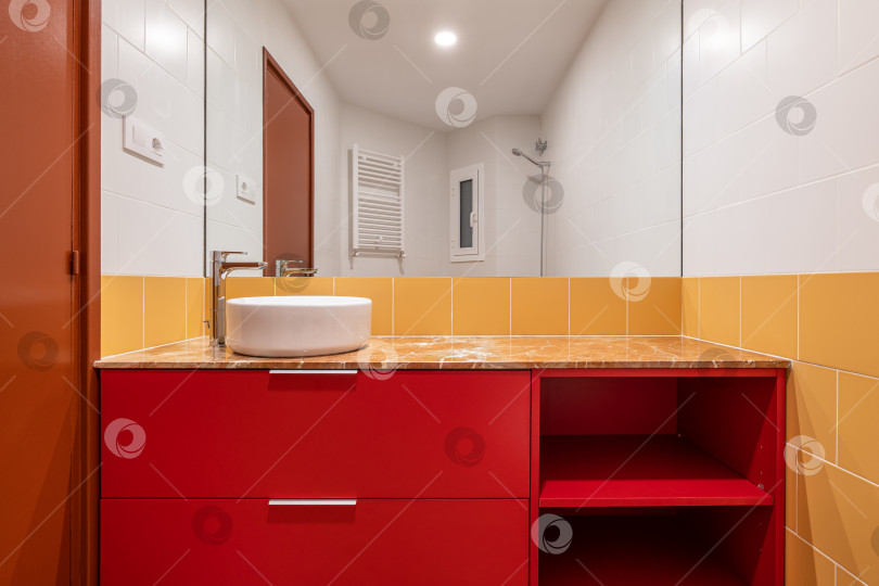 Скачать Белая раковина над головой на красном шкафчике в яркой бело-желтой ванной комнате, стены выложены белой и желтой плиткой, а пол выложен коричневой керамогранитной плиткой. Огромное настенное зеркало в ванной комнате. Коричневая дверь ванной комнаты фотосток Ozero