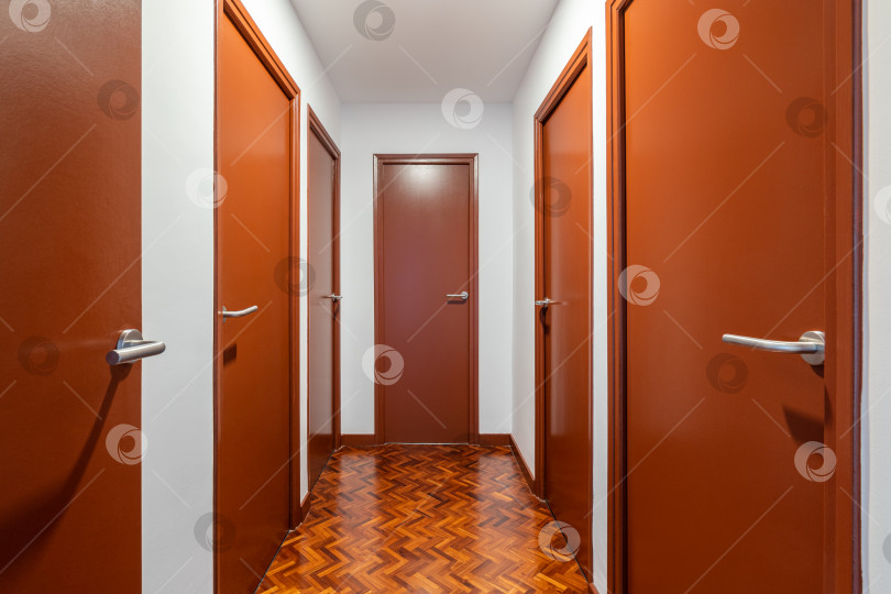 Скачать Ярко освещенный длинный коридор со множеством дверей того же темно-оранжевого цвета, ведущих в отдельные квартиры. Пол выложен паркетом с абстрактным рисунком, который соответствует цветовой гамме дверей. фотосток Ozero