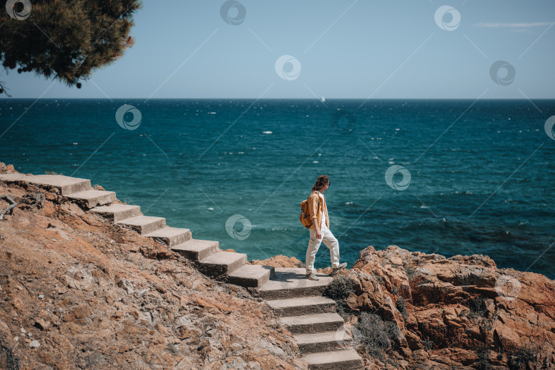 Скачать Путешественник с длинными волосами стоит на лестнице и смотрит на глубокое синее море. Концепция путешествий и пеших походов летом. фотосток Ozero