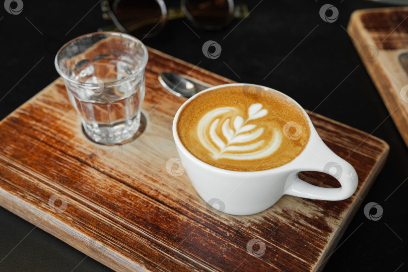 Скачать Чашка капучино с латте-артом в уютном кафе. Кофе и стакан воды на деревянной доске фотосток Ozero