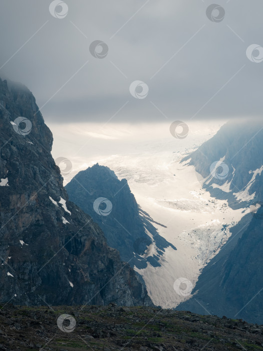 Скачать Атмосферный вертикальный пейзаж с нечеткими силуэтами острых скал и заснеженной горной вершины в низких облаках во время дождя. Впечатляющий вид на большой ледник, размытый дождевой дымкой в серых низких облаках фотосток Ozero