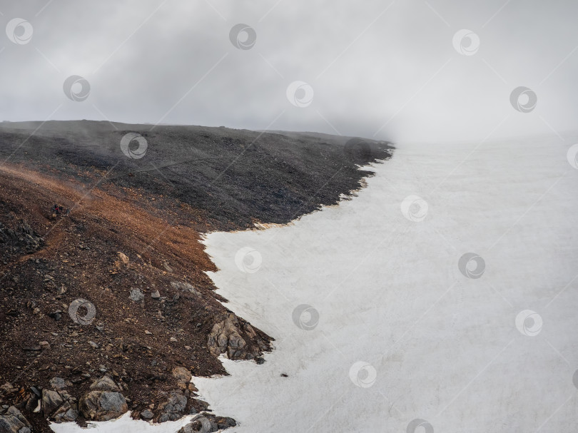 Скачать Туманный пейзаж с темным каменным склоном холма до высокой снежной горы в солнечном свете под серым облачным небом. фотосток Ozero