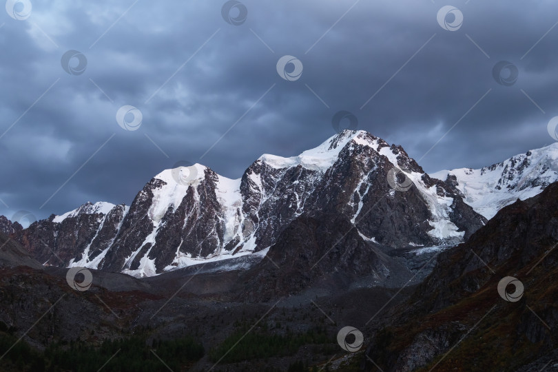 Скачать Темный атмосферный альпийский пейзаж с заснеженной горной вершиной под ночным небом. Потрясающий пейзаж с красивым остроконечным пиком, покрытым снегом, и высокой снежной горной стеной с темными низкими облаками. фотосток Ozero