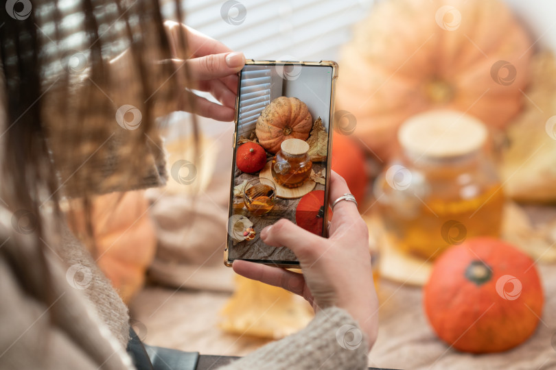 Скачать Девушка фотографирует тыквы, осенние листья и чайник на окне. Девушка фотографирует на телефон деревенскую композицию на Хэллоуин. Счастливого Дня благодарения и Хэллоуина фотосток Ozero