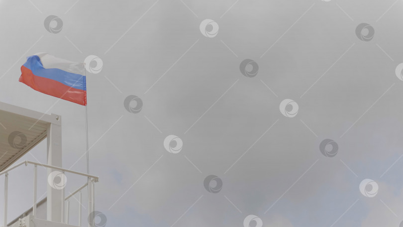 Скачать Российский флаг на флагштоке развевается на ветру на фоне голубого неба с облаками. фотосток Ozero