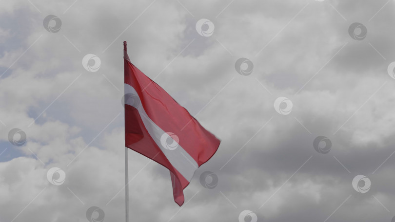 Скачать Флаг Латвии на флагштоке развевается на ветру на фоне голубого неба с облаками. фотосток Ozero