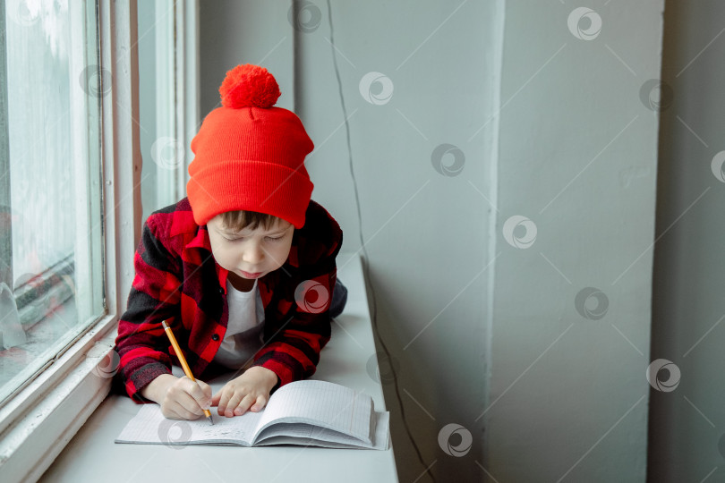 Скачать мальчик делает домашнее задание. Мальчик лежит на подоконнике и пишет в блокноте. Школьник делает домашнее задание у окна. Домашнее обучение. фотосток Ozero
