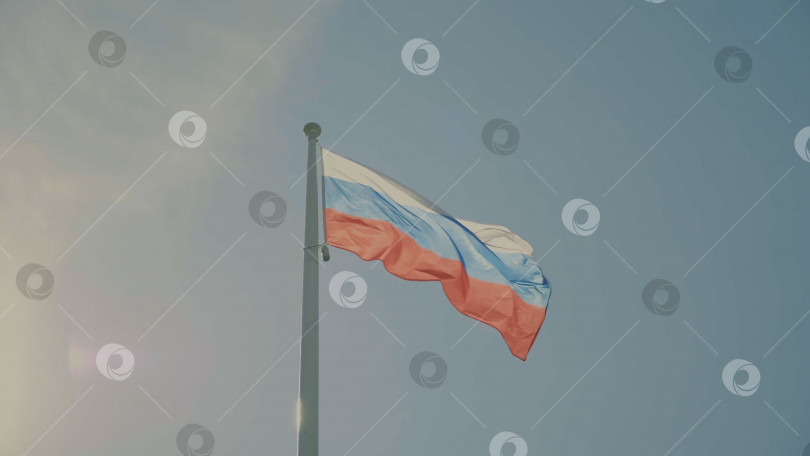 Скачать Российский флаг на флагштоке развевается на ветру на фоне голубого неба с облаками. фотосток Ozero