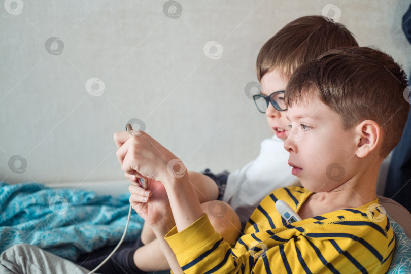 Скачать два мальчика лежат на кровати, на больничном, с градусником подмышкой и телефоном в руках. Больные дети дома фотосток Ozero