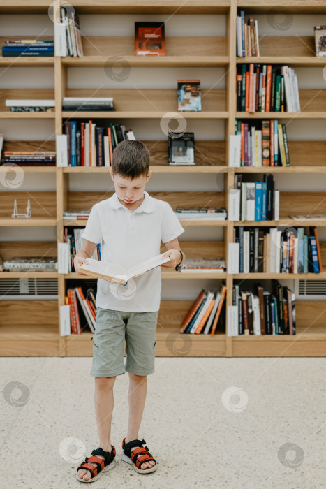 Скачать Мальчик стоит в библиотеке и читает книгу стоя. Подготовка к домашнему заданию. Мальчик любит читать. Свободное место в школе. Внеклассное обучение. фотосток Ozero