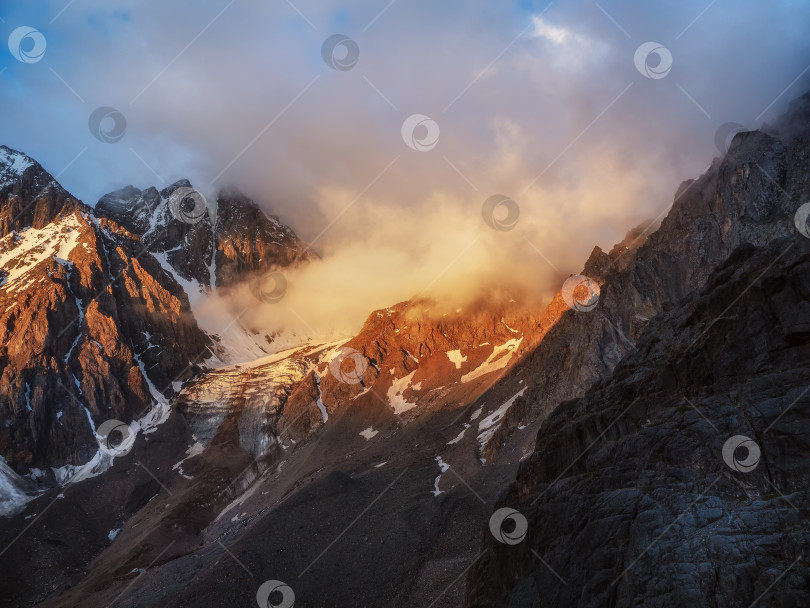Скачать Солнечный свет в горах. Большой ледник на вершине в оранжевом свете. Живописный горный пейзаж с огромным заснеженным горным хребтом, освещенным рассветным солнцем среди низких облаков. фотосток Ozero