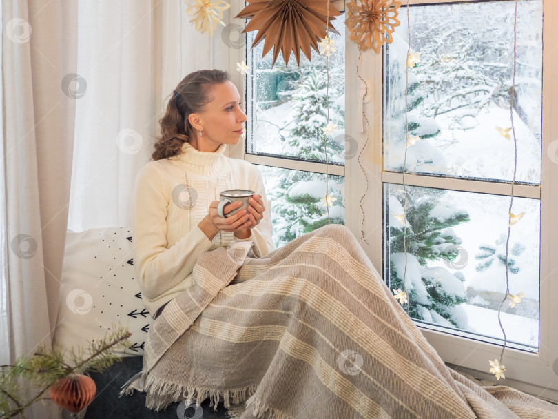 Скачать Милая женщина в уютном белом шерстяном свитере сидит у зимнего окна под теплым одеялом и держит в руках горячий кофе. Концепция домашнего уюта и комфортабельности в канун Рождества. фотосток Ozero