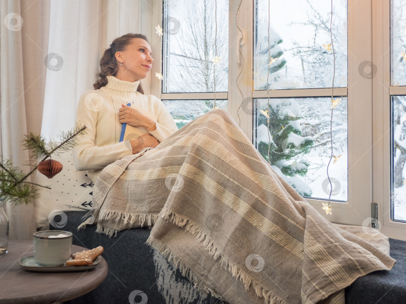 Скачать Милая женщина в уютном белом шерстяном свитере сидит у зимнего окна под теплым одеялом и прижимает к груди свою любимую книгу. Концепция домашнего комфорта и уюта в канун Рождества. фотосток Ozero