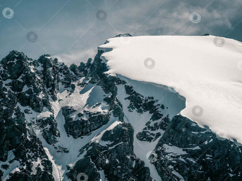 Скачать Яркий альпийский пейзаж с заснеженной вершиной горы и черными скалами в солнечном свете. Альпийский минимализм с заснеженной горой на фоне голубого неба. Минимальный вид на вершину снежной горы на очень большой высоте. фотосток Ozero