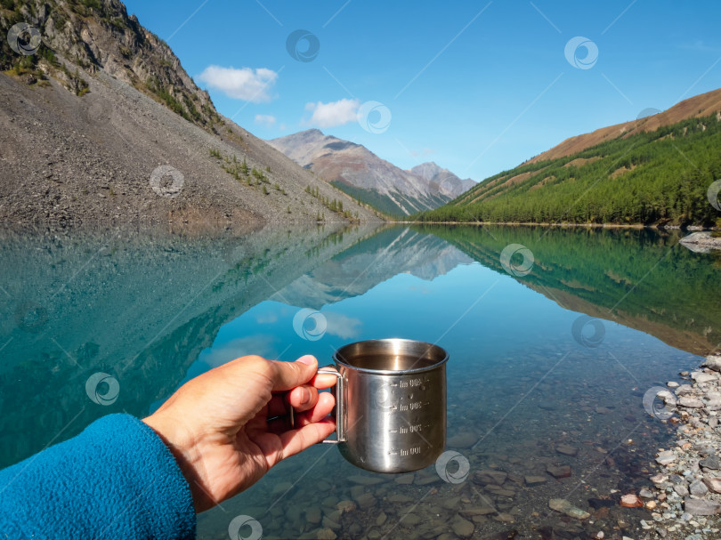 Скачать Рука, держащая походную кружку с чистой водой. Чистая питьевая вода в голубом горном озере. Чистая вода в туристическом стаканчике на фоне озера и заснеженных гор. фотосток Ozero