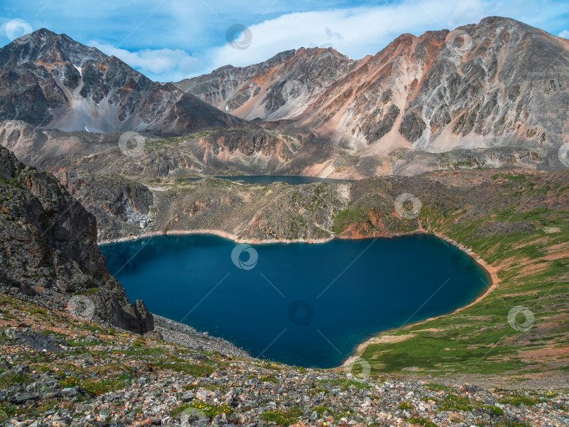 Скачать Потрясающий вид на красивое голубое горное озеро при ярком солнце. Голубое альпийское озеро в форме сердца среди залитых солнцем зеленых холмов и больших гор под солнцем в облачном небе. фотосток Ozero