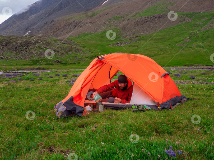 Скачать Одиночный поход. Разбив лагерь на летнем зеленом скалистом высокогорном плато. Мужчина в оранжевой палатке готовит еду. Покой и расслабление на природе. фотосток Ozero