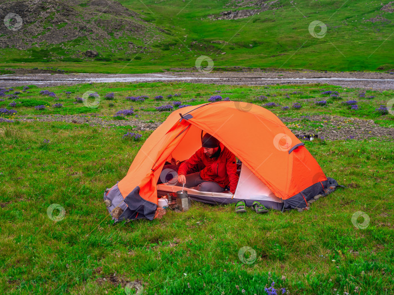 Скачать Одиночный поход. Разбив лагерь на летнем зеленом скалистом высокогорном плато. Мужчина в оранжевой палатке готовит еду. Покой и расслабление на природе. фотосток Ozero