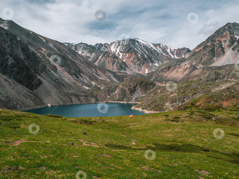 Скачать Атмосферный горный пейзаж с маленькой оранжевой палаткой рядом с альпийским озером. Потрясающие зеленые пейзажи в высокогорной долине с прозрачным горным озером с видом на заснеженные горы. фотосток Ozero