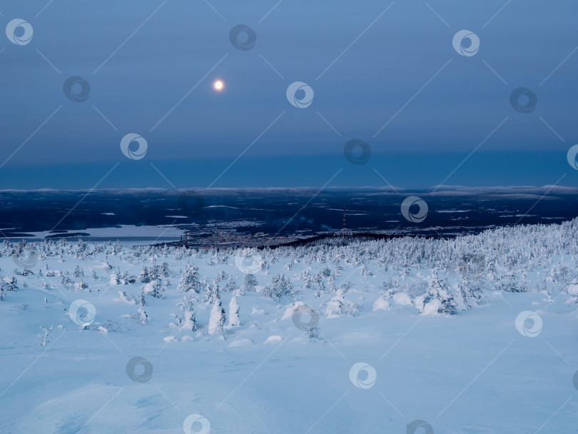 Скачать Полная луна над заснеженным склоном полярной сопки зимой с отдаленным видом на Кандалакшу, портовый город на севере России. Зимний полярный пейзаж. Холодная зимняя погода. Суровый северный климат. фотосток Ozero