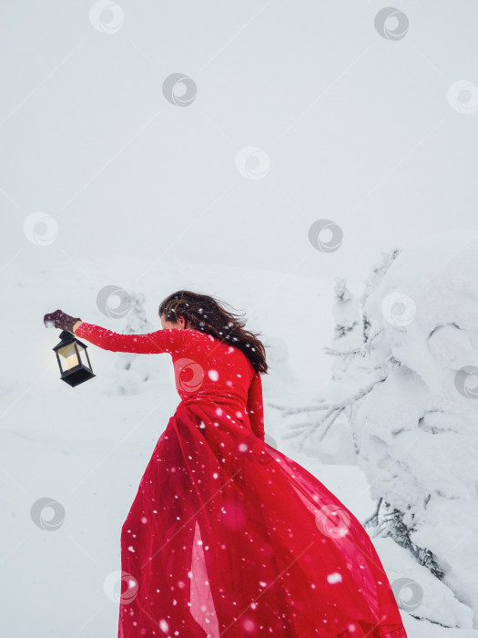 Скачать Молодая красивая женщина в длинном красном платье со старым фонарем на фоне зимнего холма и снегопада. Сказочная девочка на фоне полярного зимнего пейзажа. Рыжая ведьма в снегу. Вертикальный вид. фотосток Ozero