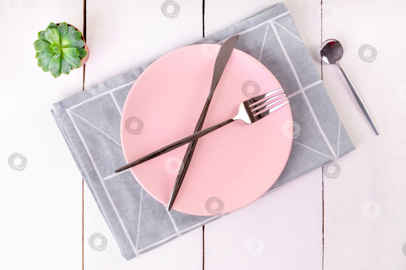 Скачать Сервировочная пустая розовая тарелка со скрещенными ножом и вилками на сложенной льняной салфетке с геометрическим рисунком фотосток Ozero